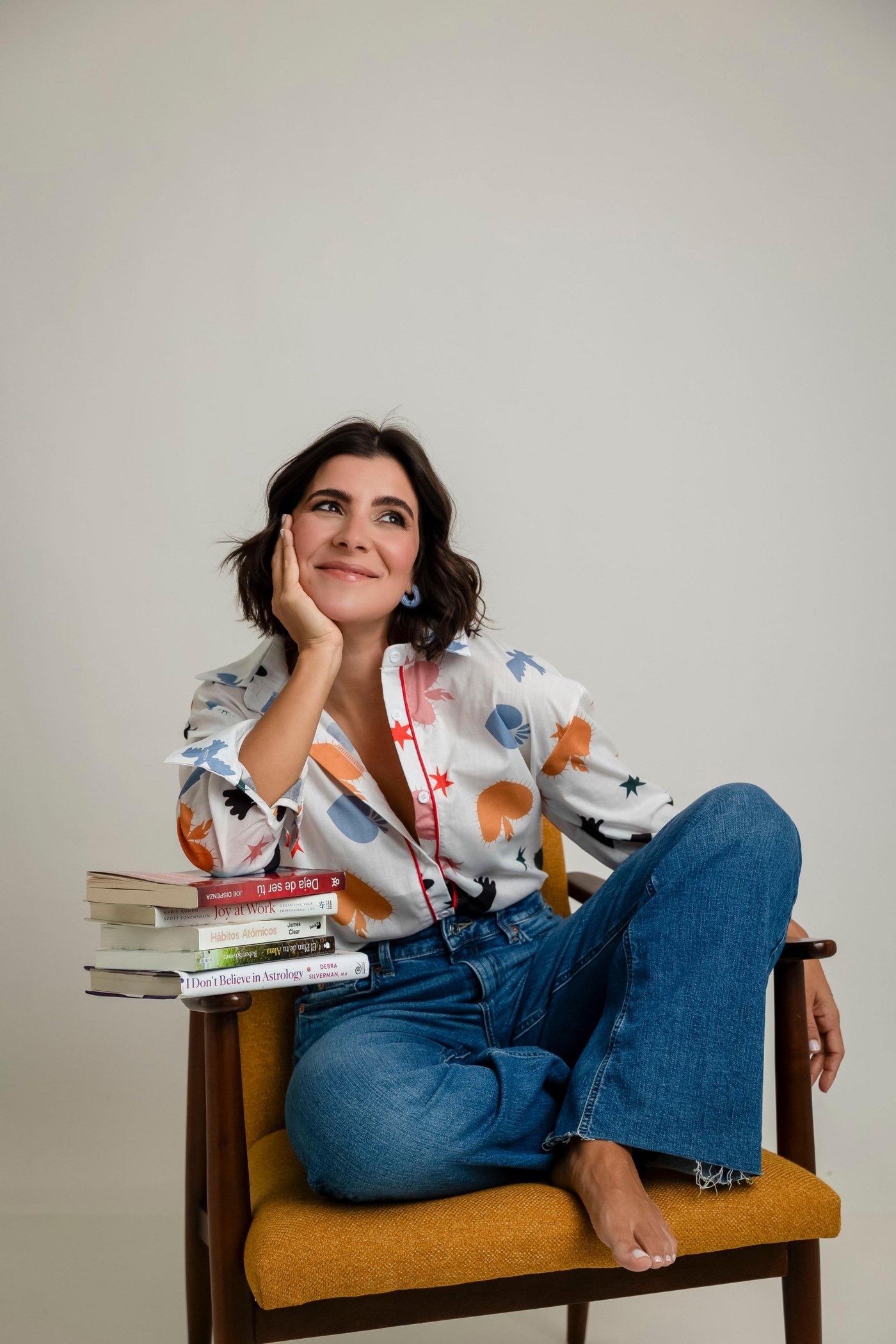 Cristina Badell sentada en una silla color mostaza, sonriendo con la mano en la mejilla, junto a una pila de libros.