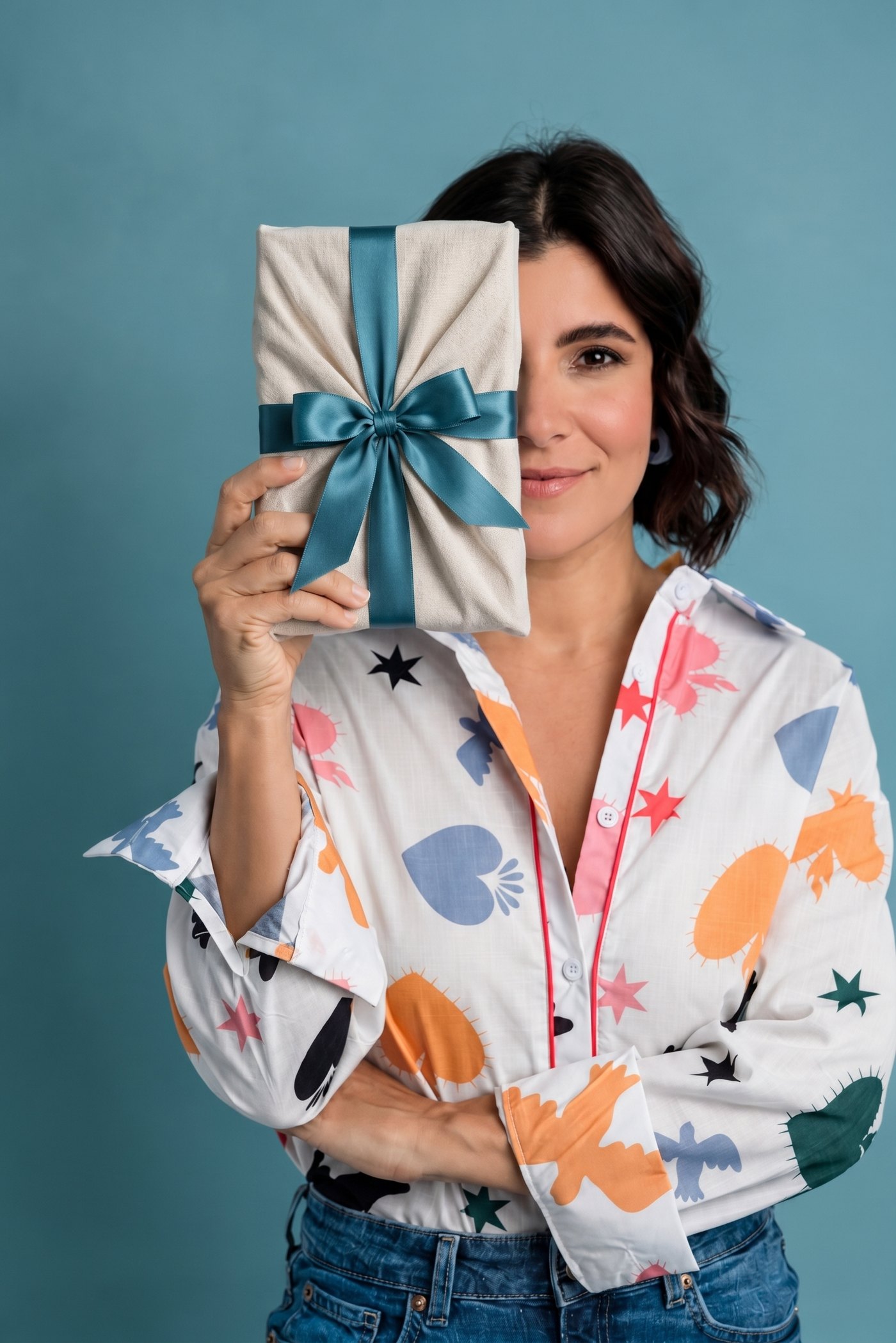 Cristina Badell sonríe sosteniendo frente a su rostro el libro envuelto como un regalo, con tela cruda y un lazo teal, sobre un fondo azul verdoso.